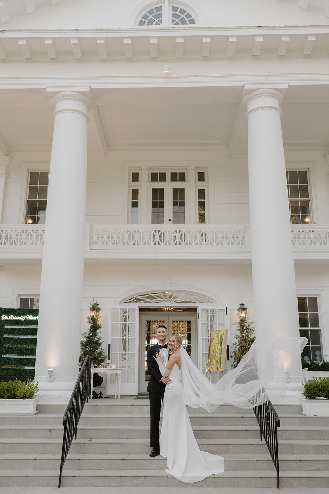 Bride shows off her ring while posing with the groom on the steps, her veil blowing dramatically in the wind.